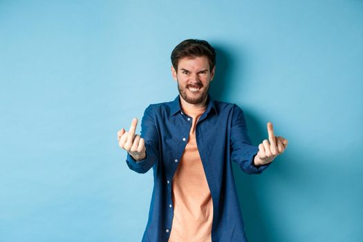 Angry caucasian man say fuck you and showing middle fingers, make rude gesture with pissed-off face, standing annoyed on blue background.