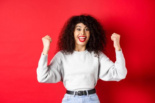 Cheerful lucky woman with curly hair, winning prize and scream yes with joy, raising hands up and celebrating, triumphing and cheering, standing on red background.