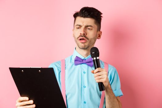 Holidays concept. Serious-looking man reading script from clipboard, holding microphone, entertain people on festive event, standing on pink background.