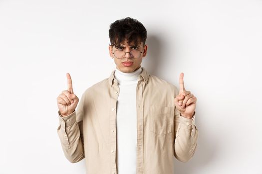 Angry and displeased young man in glasses frowning, looking disappointed and pointing fingers up, standing on white background.