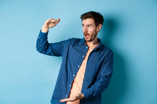 Impressed caucasian guy staring at empty space with big object, shaping large size, standing on blue background. Copy space