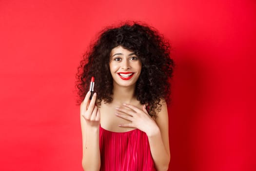 Beauty and make up concept. Beautiful female model with curly hair, wearing evening dress, showing red lipstick and smiling, standing on white background.