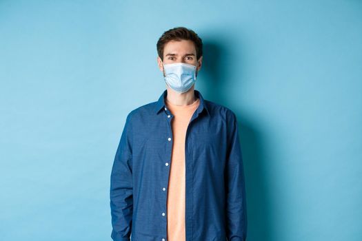 Covid-19 and healthcare concept. Smiling young man in medical mask looking healthy and happy, standing on blue background.