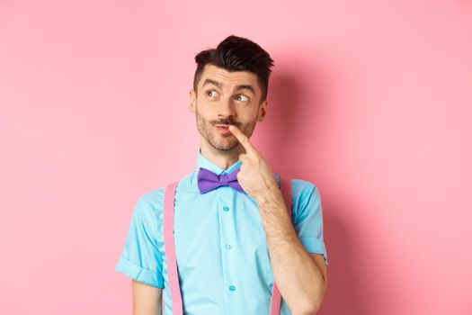 Image of handsome funny man in bow-tie looking thoughtful left and touching lip, thinking what to choose, making decision, standing over pink background.