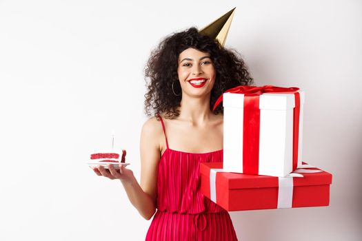 Happy birthday girl in red dress, celebrating and holding gifts with bday cake, standing on white background.