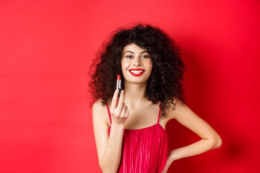 Fashionable woman with curly hair, showing red lipstick and smiling, recommend cosmetic, standing on white background.