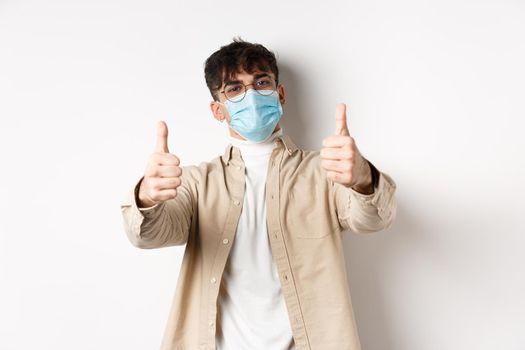 Coronavirus, health and real people concept. Smiling guy in medical mask showing thumbs up, wearing glasses, standing on white background.