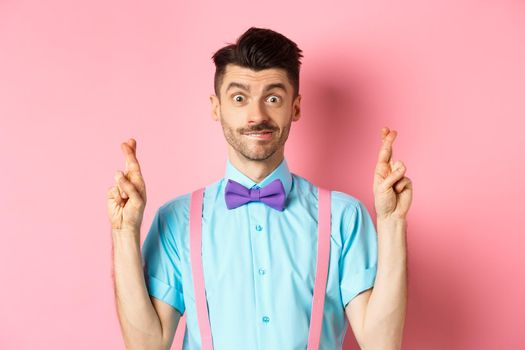 Hopeful young man making wish, holding fingers crossed and smiling, waiting for results with optimism, standing over pink background.