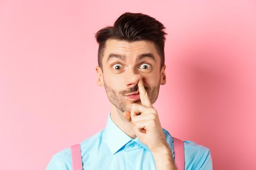 Close-up of funny caucasian man picking nose, staring silly at camera, standing on pink background. Copy space