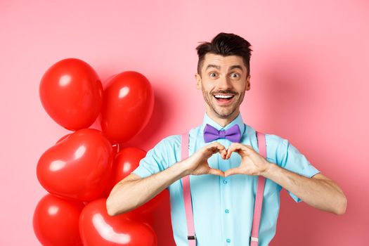Valentines day concept. Romantic guy looking happy and smiling, showing heart gesture to lover on special anniversary, standing on pink background.