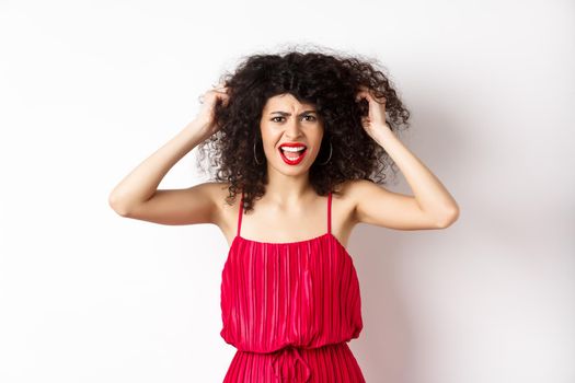 Frustrated curly woman in red dress, frowning and screaming angry, pull out hair and shout at camera, standing on white background.