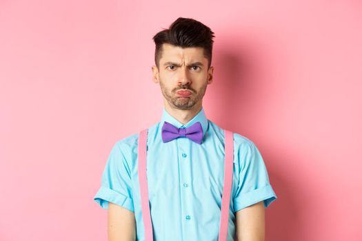 Sad and offended young man, frowning and pucker lips, looking angry at someone, standing disappointed against pink background in bow-tie.