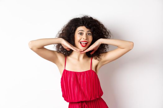 Beauty. Happy elegant woman with curly hair and makeup, wearing red dress, showing her face and smiling excited, standing against studio background.