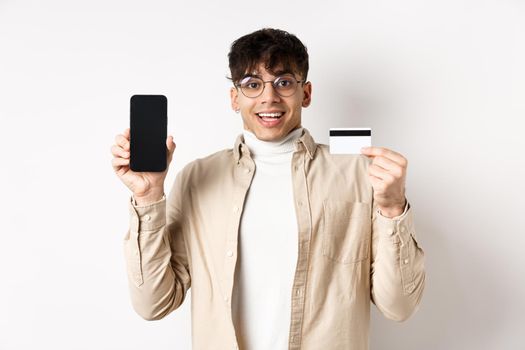 Online shopping. Surprised and happy young man showing credit card and mobile phone screen, standing on white background.