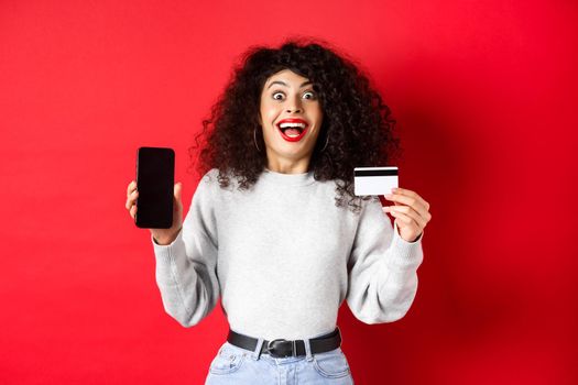 Online shopping. Happy young woman showing plastic credit card and empty phone screen, announce promo offer, standing on red background.