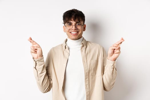 Handsome and positive young man smiling, making wish with crossed fingers and happy face, hope for dream to come true, standing on white background.