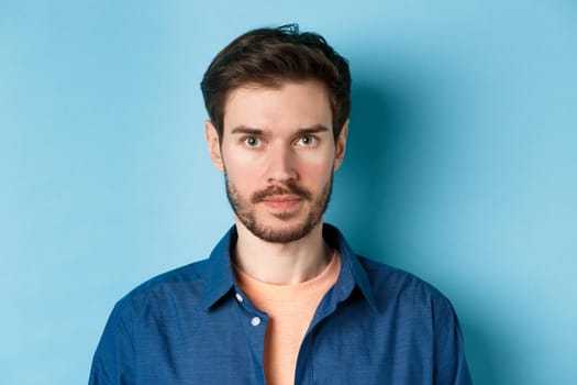 Close-up of young man with beard looking serious at camera, standing in casual shirt on blue background.