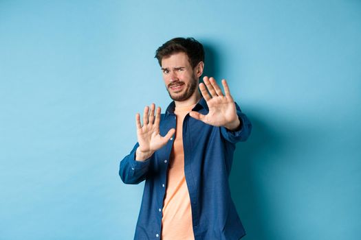 Guy cringe from something disgusting or embarrasssing, stretching out hands and asking to stop, grimacing displeased, standing on blue background.