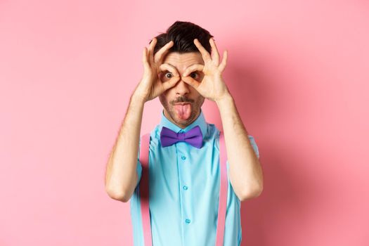 Funny and silly young man in bow-tie and suspenders, looking through hand-binoculars and showing tongue at camera, standing cheerful on pink background.