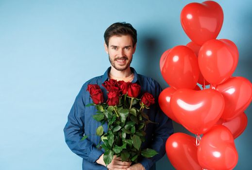 Handsome romantic man holding red roses and smiling, standing near heart balloons over blue background.
