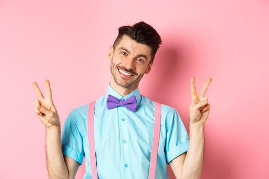 Romantic guy in bow-tie and suspenders showing peace signs and smiling at camera, wearing clothes for date, standing on pink background.