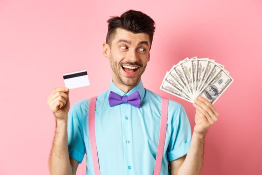 Image of smiling young man in bow-tie holding plastic credit card and looking happy at money, choosing cash, standing over pink background.