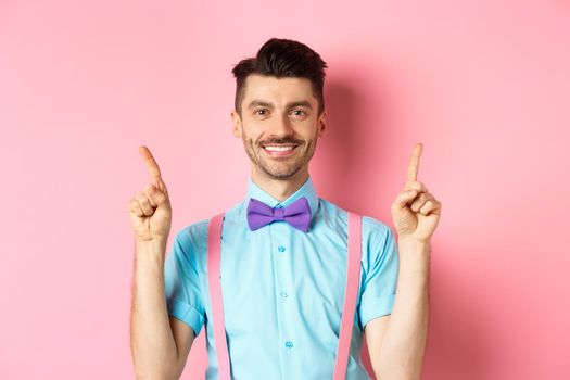 Cheerful handsome guy smiling at camera, pointing fingers up, showing advertisement on top of pink background.