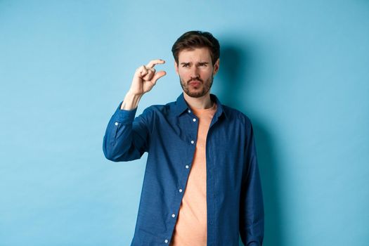 Confused guy showing small size and looking doubtful or skeptical, standing over blue background. Copy space
