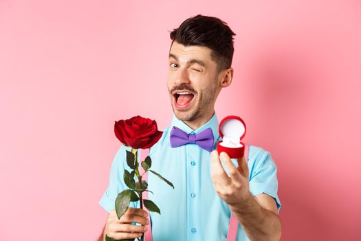 Valentines day. Funny guy making proposal, winking and saying marry me, showing engagement ring with red rose, standing over pink background.