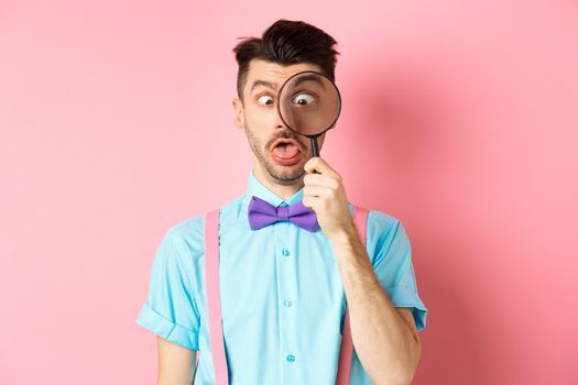 Funny man in bow-tie look through magnifying glass, squinting and making silly faces, standing on pink background.