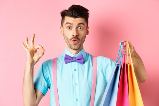 Funny guy in bow-tie showing OK sign and shopping bags, praising good deal in shops, standing over pink background.