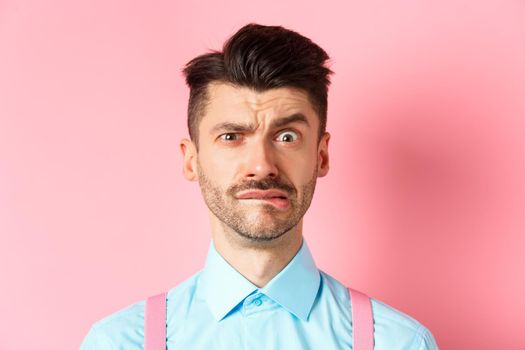Close-up of anxious young guy with moustache, biting lip and frowning, looking worried over small mistake, standing on pink background.