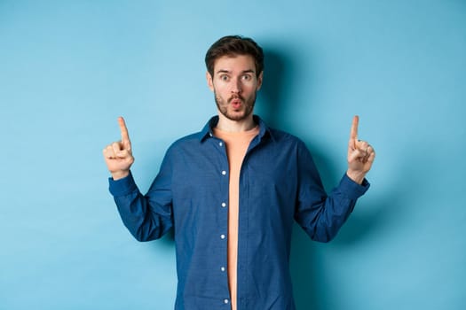Surprised young man say wow, checking out advertisement, pointing fingers up and looking impressed, standing on blue background.