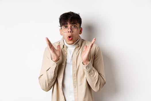 Portrait of surprised hipster guy in glasses checking out awesome promo, say wow and gasping amazed, raising hands up with disbelief, standing on white background.