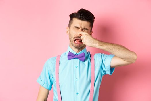 Image of man crying from disgusting smell, shut his nose from awful stink, standing in bow-tie and suspenders on pink background.