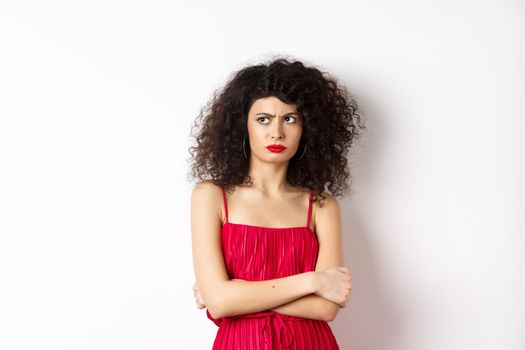 Suspicious and reluctant elegant woman, wearing red dress and frowning, looking left with disbelief, standing on white background.