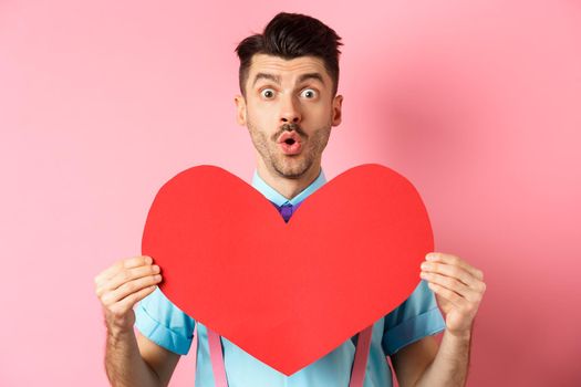 Valentines day concept. Amused young man with bow-tie and moustache, showing heart cutout and looking for love, standing on pink romantic background.