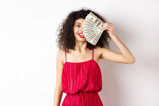 Happy curly woman winning money, holding dollar bills on half of face, smiling excited, standing in red dress on white background.