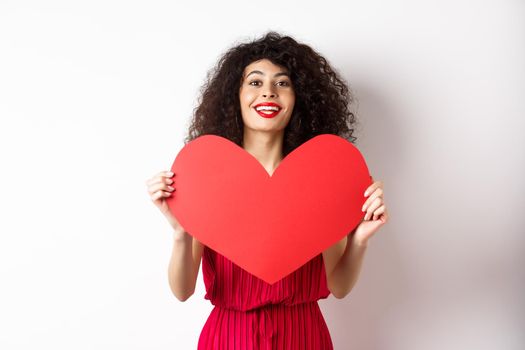 Romantic woman in dress showing big red heart, falling in love, smiling happy at camera, white background.