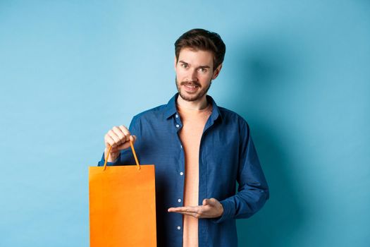 Handsome caucasian man showing orange shopping bag and smiling, purchased gift, standing on blue background. Copy space