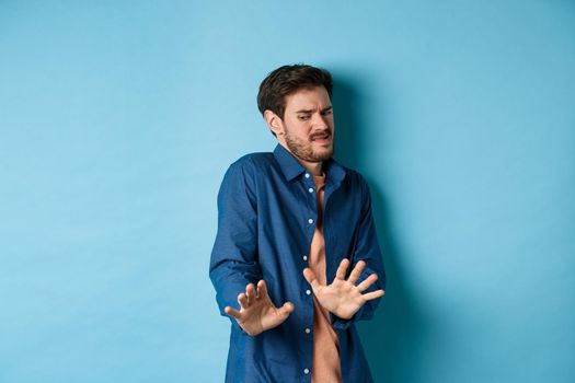 Image of man cringe and look at something disgusting, step back from awful thing and block it with hands, standing on blue background. Copy space