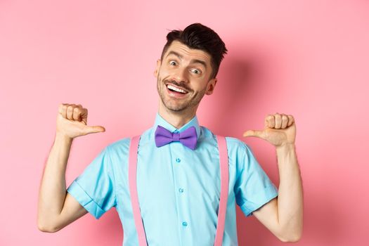 Cheerful smiling man with moustache and bow-tie, pointing at himself and self-promoting, standing happy over pink background in suspenders.