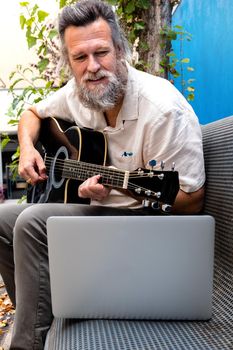 Mature caucasian man taking online guitar lessons using laptop sitting on a bench. Vertical image. Leisure activities concept. E-learning concept.