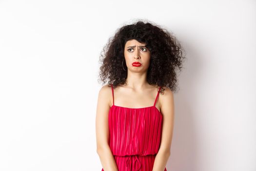 Sad and gloomy young woman with curly hair and makeup, wearing red dress, sobbing and looking jealous or upset aside, see something unfair, standing on white background.