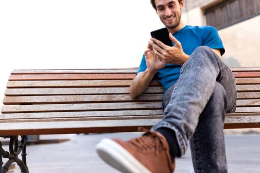 Young happy caucasian man relaxing sitting on a bench using cellphone. Selective focus on hand. Copy space. Social media, technology and lifestyle concepts.