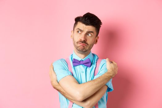 Timid guy feeling offended and lonely, embracing body, comforting himself and looking left upset, standing on pink background.