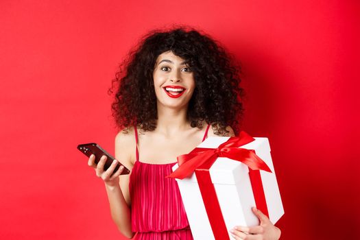 Online shopping and Valentines day. Beautiful young woman holding smartphone and lovers gift, looking surprised and happy at camera, red background.