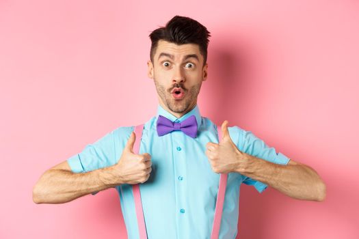 Impressed young guy in suspenders and bow-tie showing thumbs up and saying wow, recommending awesome product, praising quality, standing over pink background.