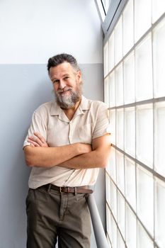 Mature caucasian man with beard looking at camera with arms crossed next to window. Vertical image. Portrait. Lifestyle concept.
