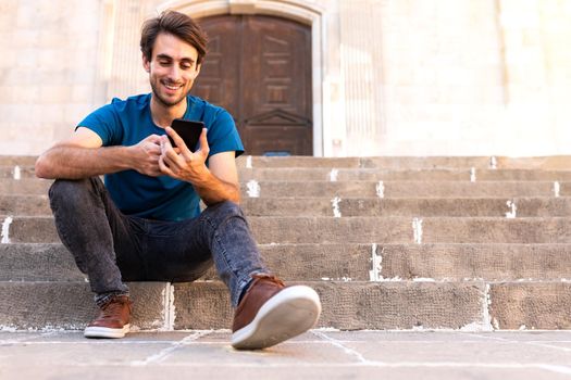 Smiling young caucasian man sitting on stairs using mobile phone to send text message. Copy space. Lifestyle and social media concept.